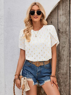 Woman wearing a white blouse with gold polka dots and denim shorts against a wooden wall.